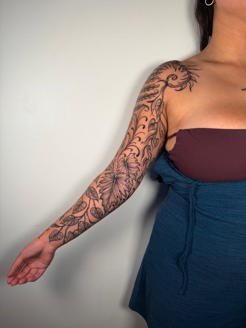 A woman's upper arm and shoulder featuring a large black floral and scroll tattoo, shown against a plain light gray wall. She is wearing a dark brown strapless top and a blue skirt.