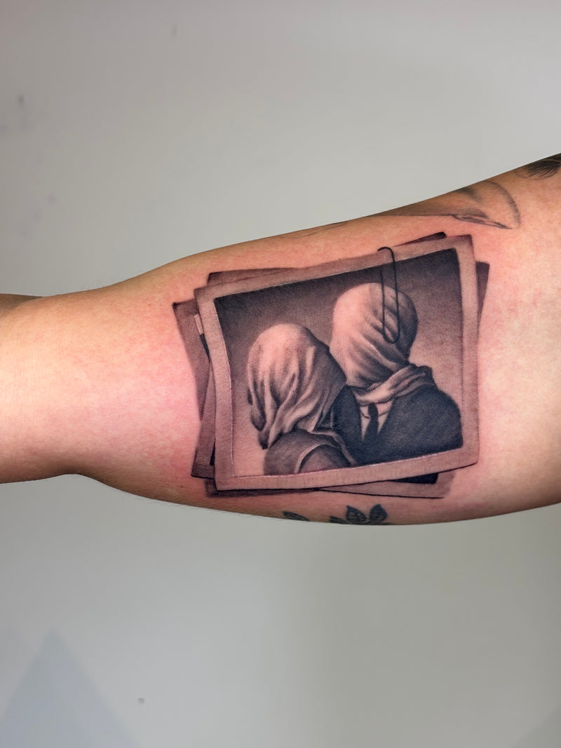A close-up of a person's forearm featuring a realistic black and gray tattoo of two figures in hooded clothing, styled inside overlapping tilted photo frames.