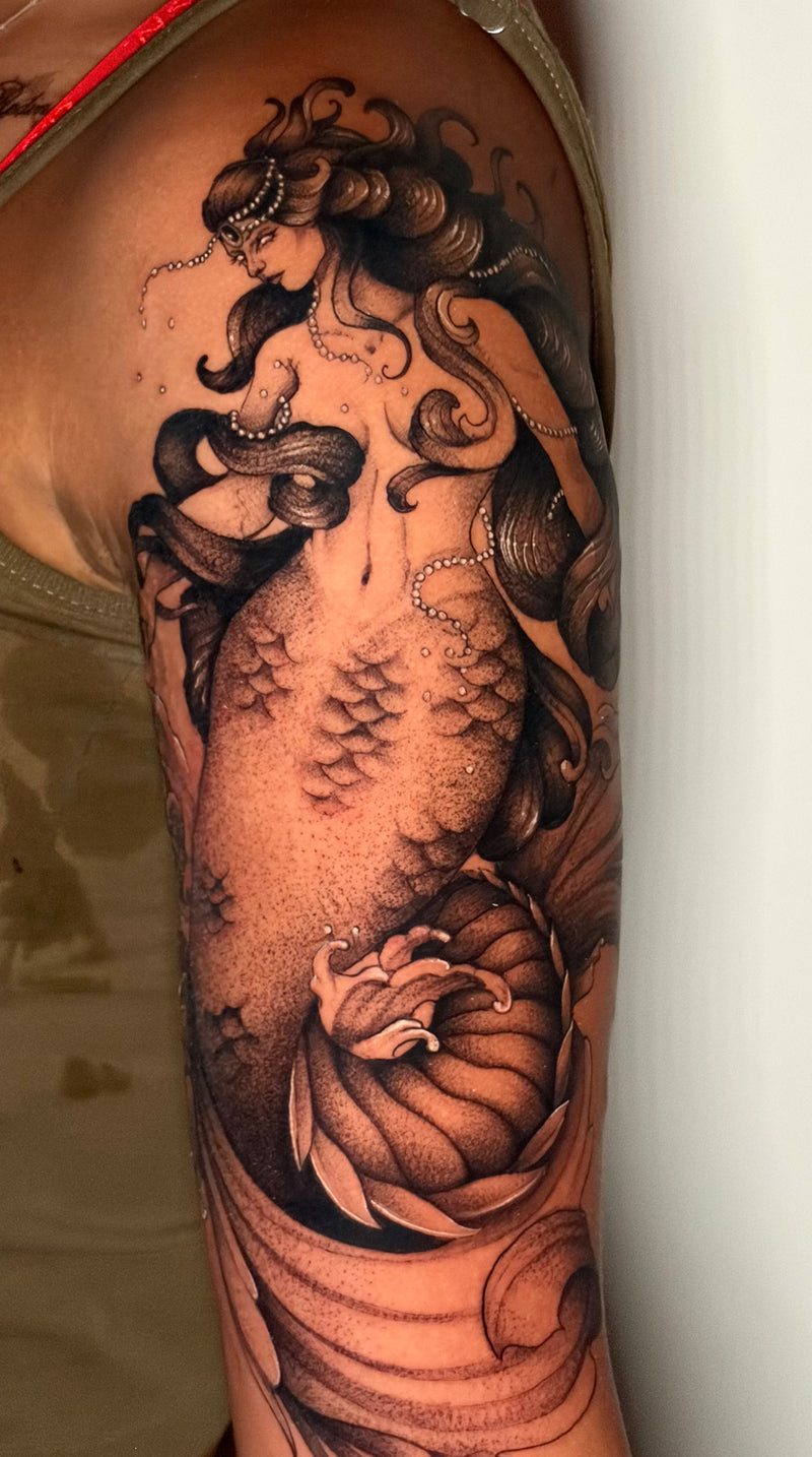 A large black and gray tattoo of a mermaid with flowing hair and a floral motif on a person's upper arm.
