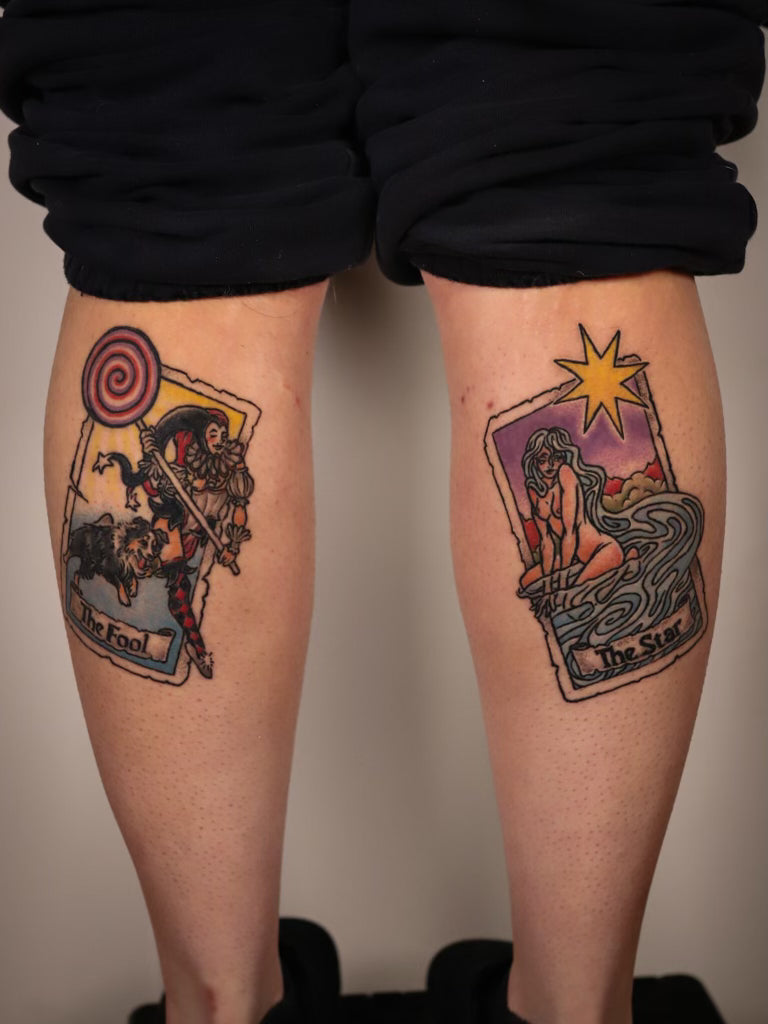 A person's legs from the back, showing two colorful tarot card tattoos on the upper calves. The left tattoo depicts 'The Fool' with a red and yellow spiral, and the right tattoo depicts 'The Star' with a yellow star and purple sky.