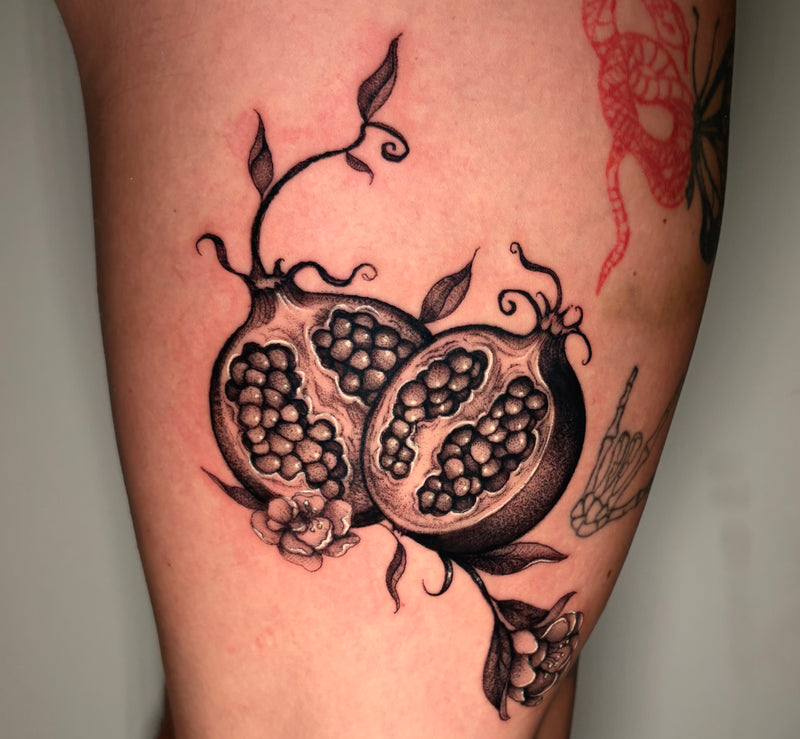 A close-up of a tattoo on skin showing two detailed pomegranate halves with black, gray, and white shading, small leaves, and flowers. Partial red and green tattoos are visible on adjacent skin.