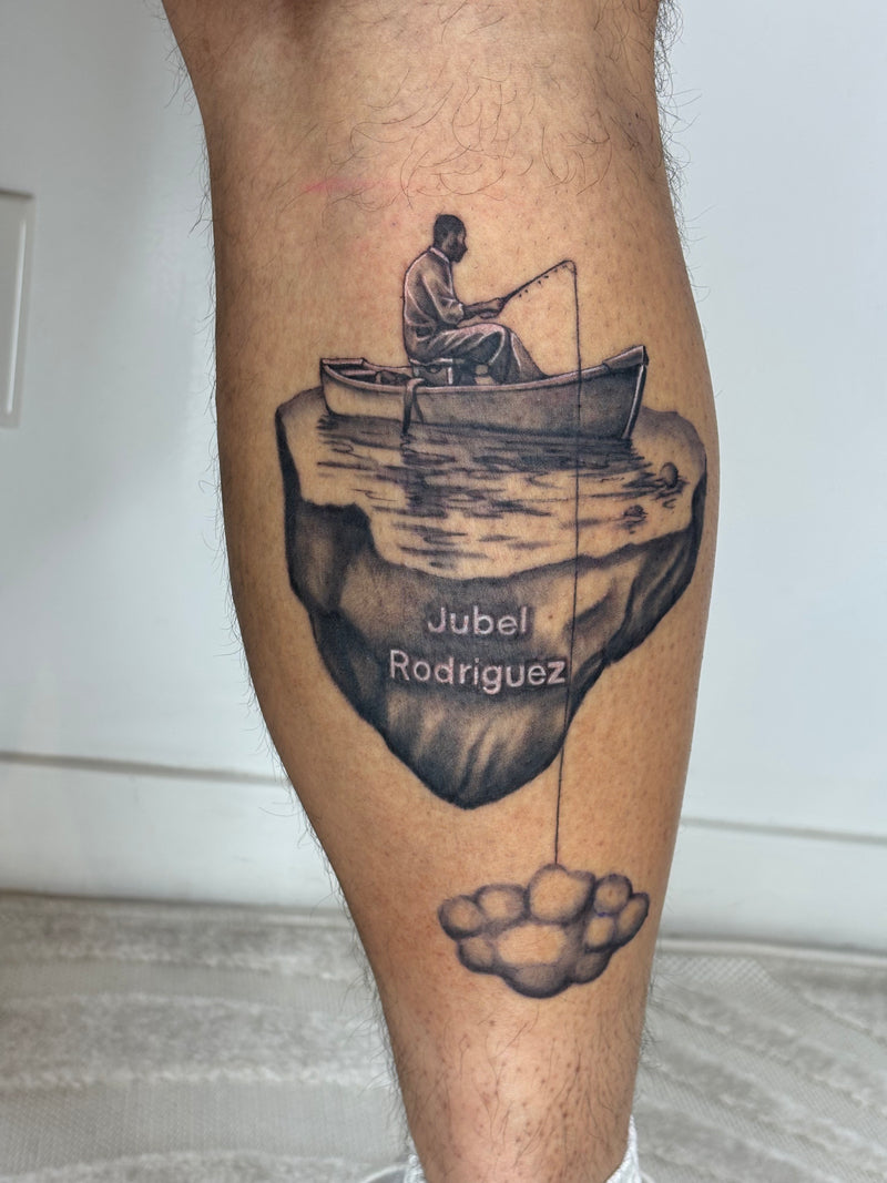 A black and gray temporary tattoo applied to a person's calf, featuring a man fishing from a cliff edge with the text 'Jubel Rodriguez' and a cloud below.