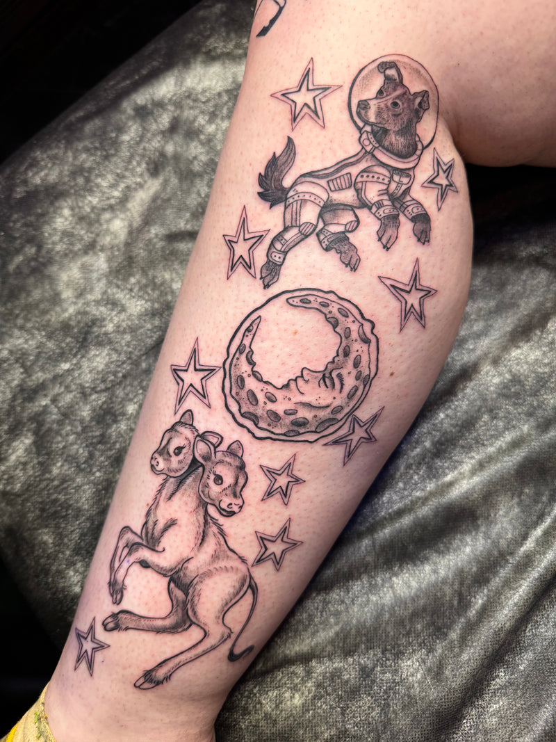 A close-up of a person's leg featuring a detailed black and gray tattoo design with a cow, stars, a crescent moon, and a dog astronaut floating in space, resting against a dark textured surface.
