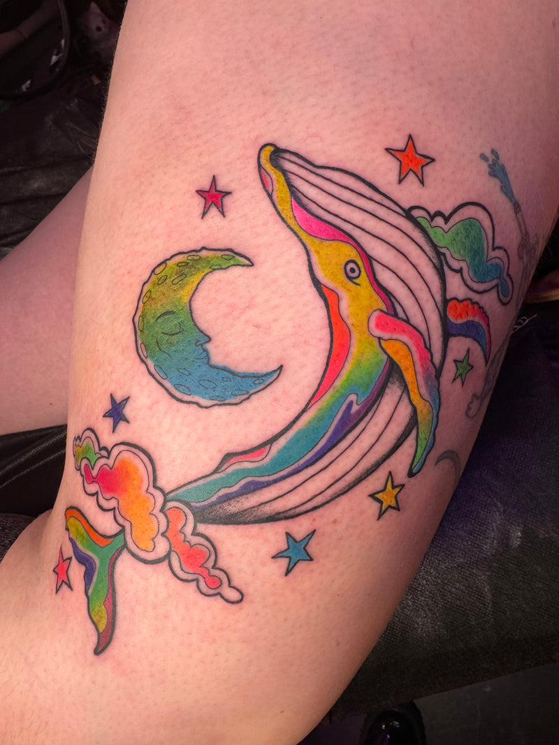 A colorful tattoo of a whale jumping over a crescent moon, surrounded by stars and clouds, applied to a person's upper arm.