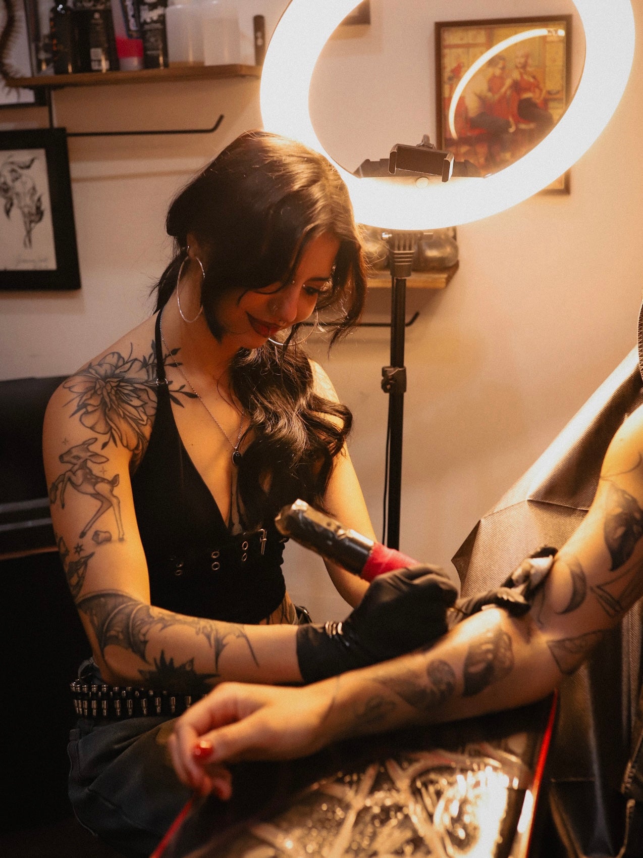 A tattoo artist with arm tattoos works on a client's arm under a bright ring light in a tattoo studio.