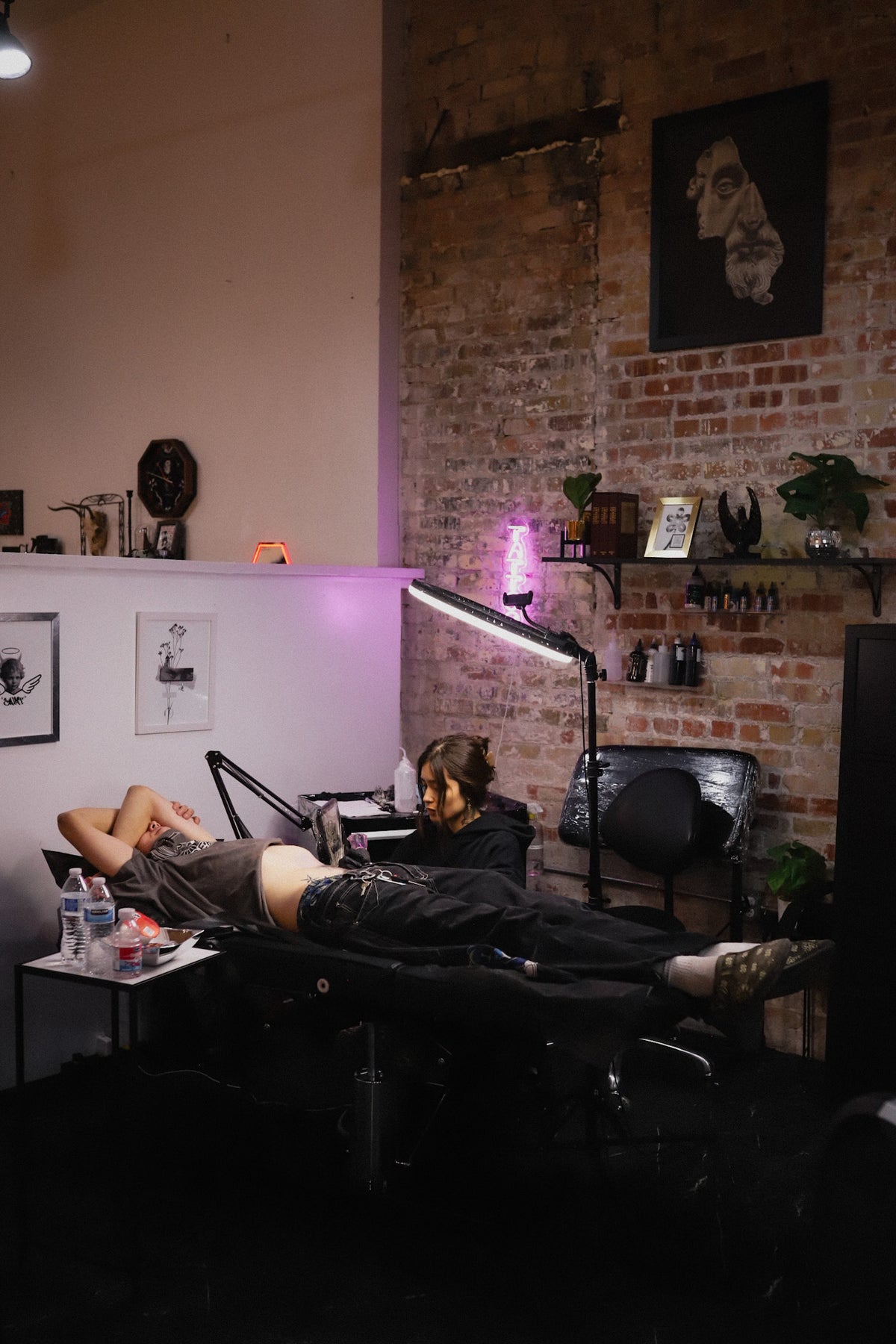 A tattoo artist sits beside a reclining client on a tattoo table in a studio with exposed brick walls, shelves, and purple ambient lighting.