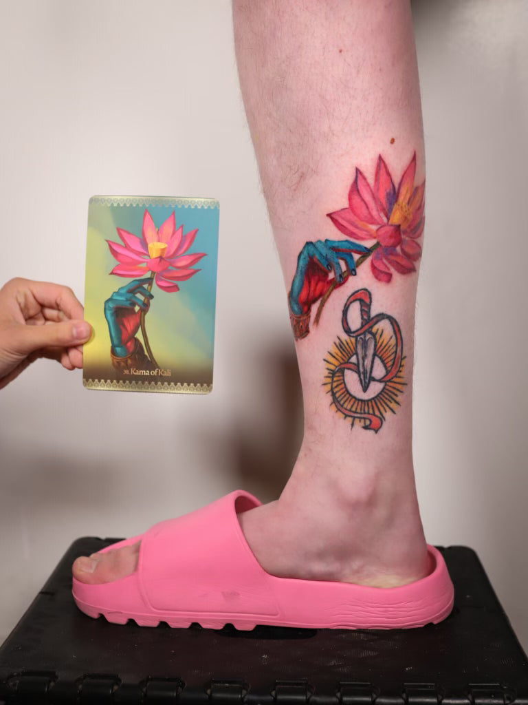 A person's lower leg and foot wearing a bright pink slide sandal, showing colorful lotus flower and mandala-style tattoos on the calf. A hand holds a matching tattoo card next to the leg against a plain indoor background.