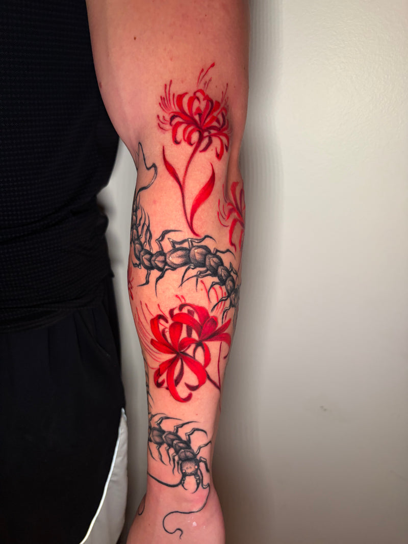 A close-up of a person's forearm featuring a tattoo design with two bright red spider lilies and black centipede illustrations on the upper arm and forearm.