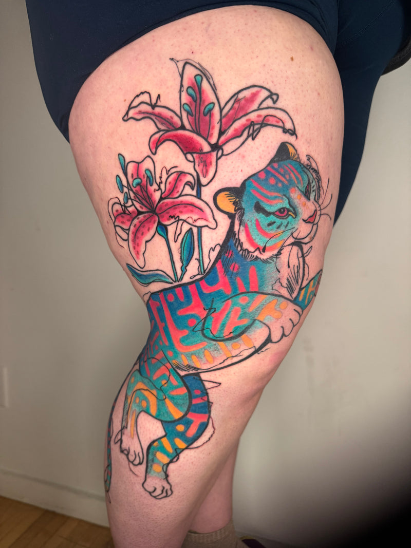 A colorful tattoo of a stylized tiger and pink lilies on a person's upper thigh, shown against a plain indoor wall.