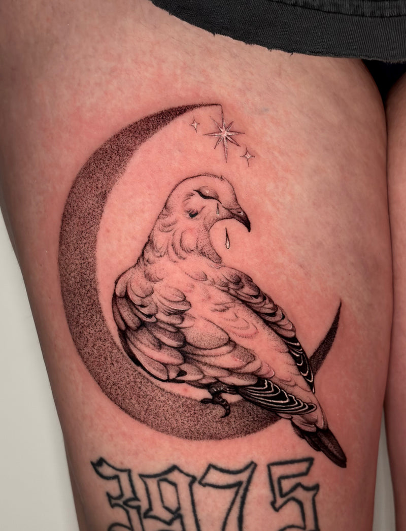 A close-up of a freshly tattooed thigh showing a detailed black and gray ink design of a bird perched on a crescent moon with stars, above the numbers 3975.