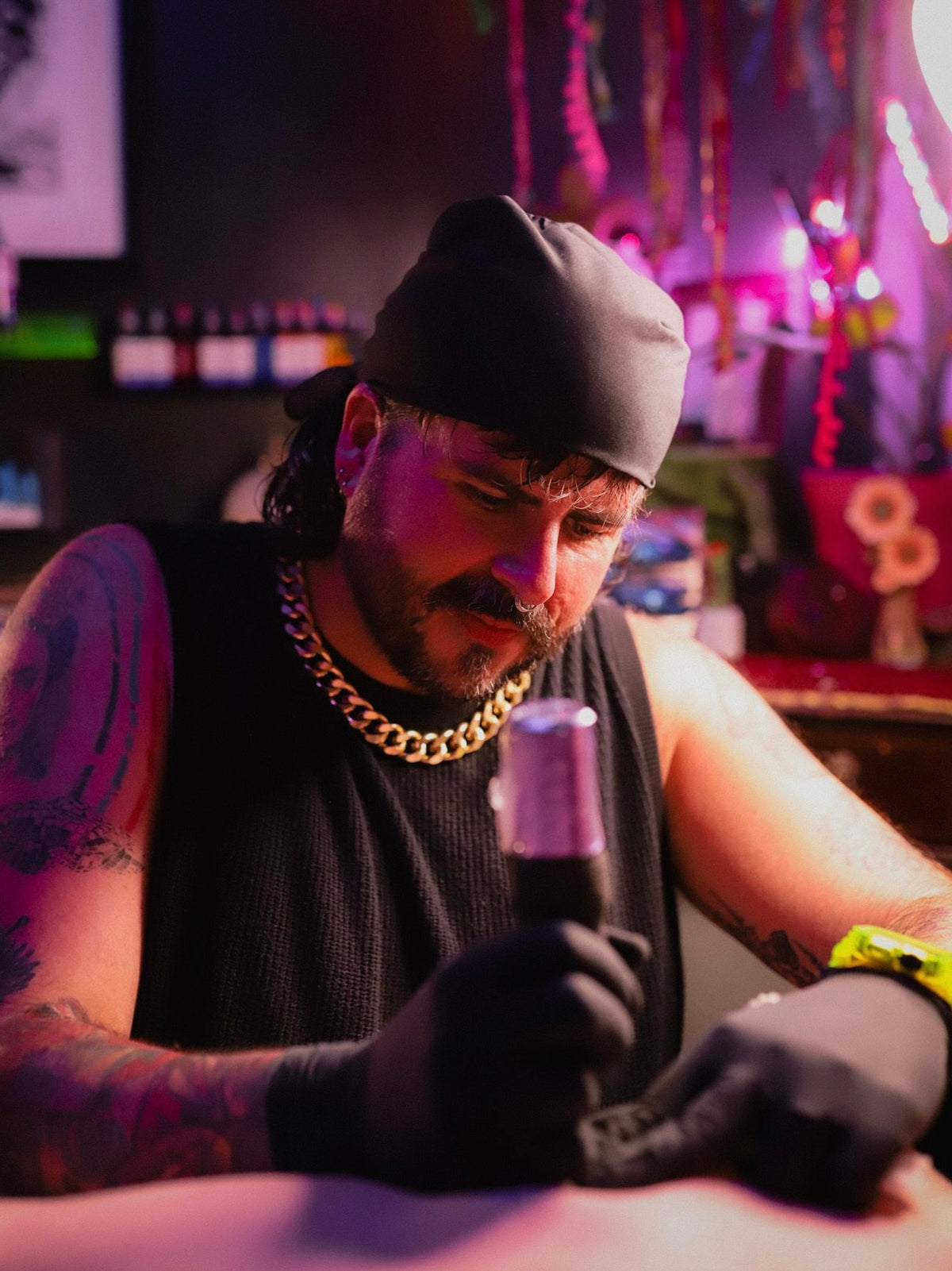 A tattoo artist wearing a black beanie, black sleeveless shirt, and black gloves holds a tattoo machine over a client's shoulder in a dimly lit studio with pink and purple ambient lighting.
