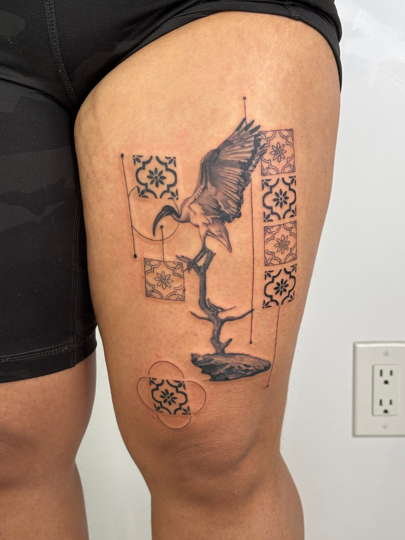 A black and gray tattoo of a heron perched on a branch, framed by decorative tile patterns, shown on a person's upper thigh.