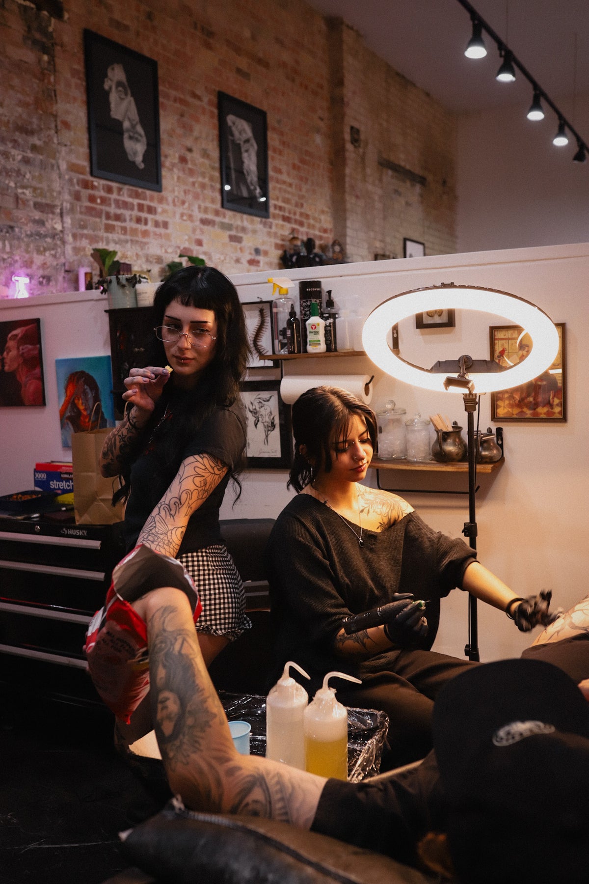 A tattoo artist with long dark hair and arm tattoos is seated beside a client receiving a tattoo in a tattoo studio. The client lies on a tattoo chair with their arm extended, wearing a black sleeveless top.