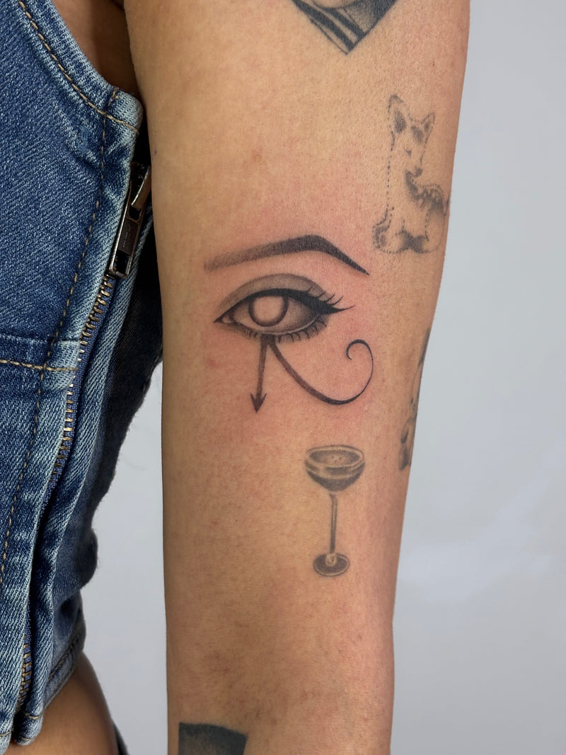 A close-up of a person's forearm featuring a black ink tattoo of the Egyptian Eye of Horus with a teardrop, accompanied by a small cocktail glass and a cat motif. The person is wearing blue denim shorts.