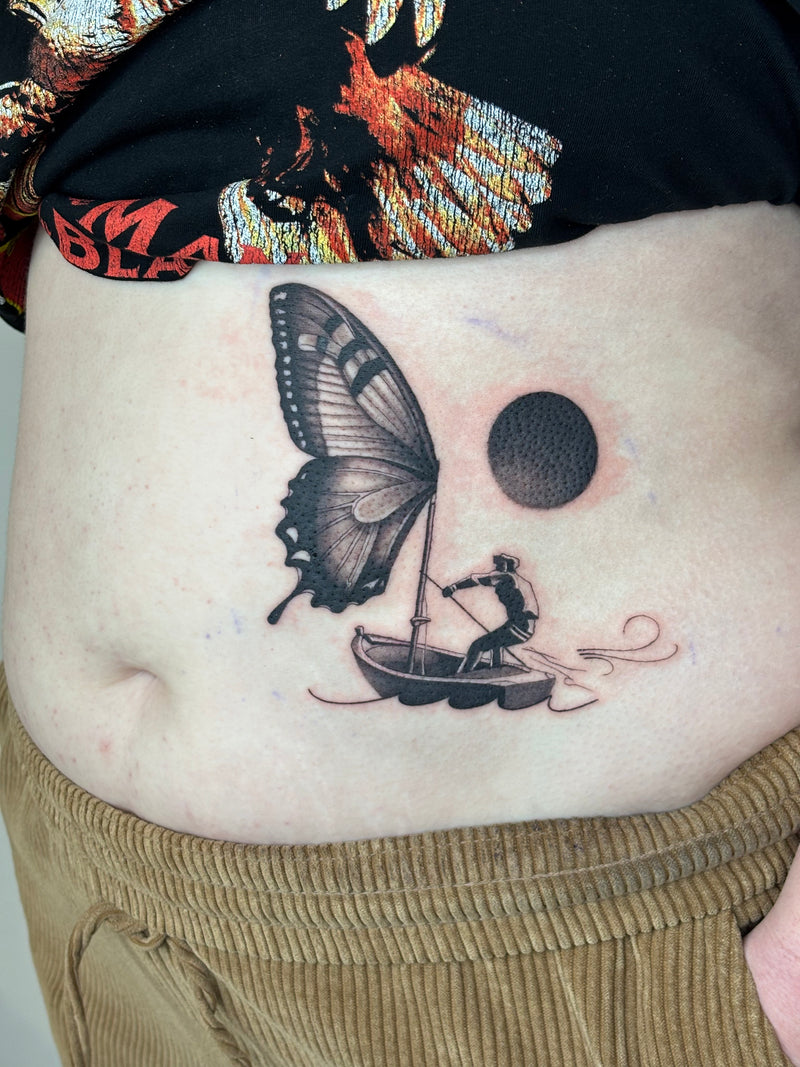 A close-up of a person's lower abdomen showing a freshly tattooed design. The black ink tattoo depicts a figure rowing a small boat under a large butterfly wing, with a dark circular sun or moon in the sky.