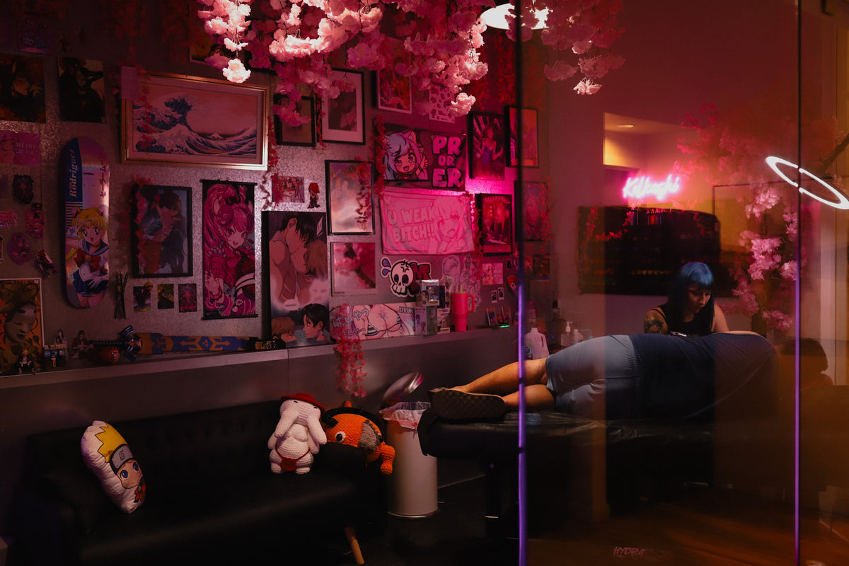 A dimly lit, highly decorated tattoo studio or lounge space with a person reclining on a table. The room is illuminated by strong pink and red ambient lighting, decorated with cherry blossom tree lights, framed anime-style posters on the wall, and plush toys on a black couch.