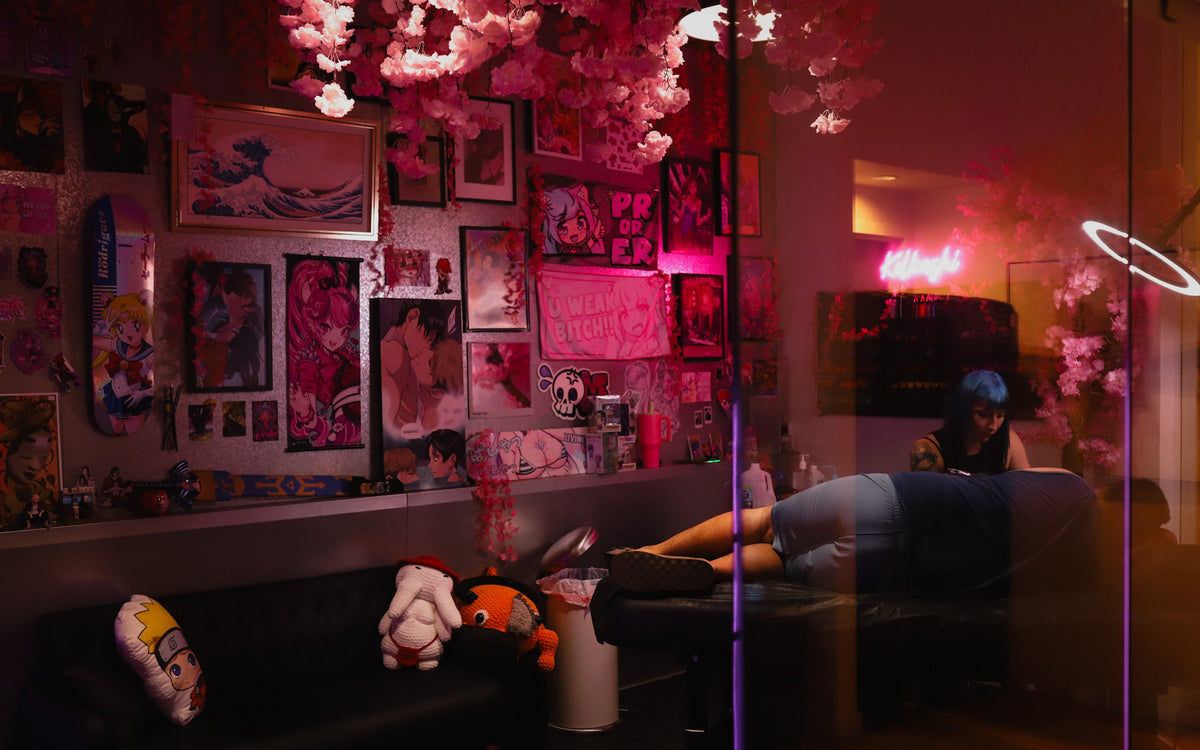 A dimly lit, highly decorated room with pink cherry blossom decorations hanging overhead, a wall covered in framed art and posters, and a person reclining on a massage table. The scene is dominated by pink and red ambient lighting.