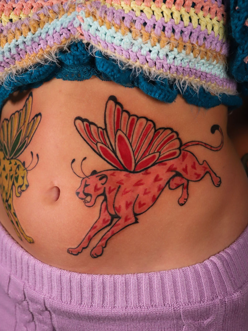 A temporary tattoo of a red and orange cheetah with a butterfly wing, applied to a person's stomach, with a colorful striped knit top visible above it.