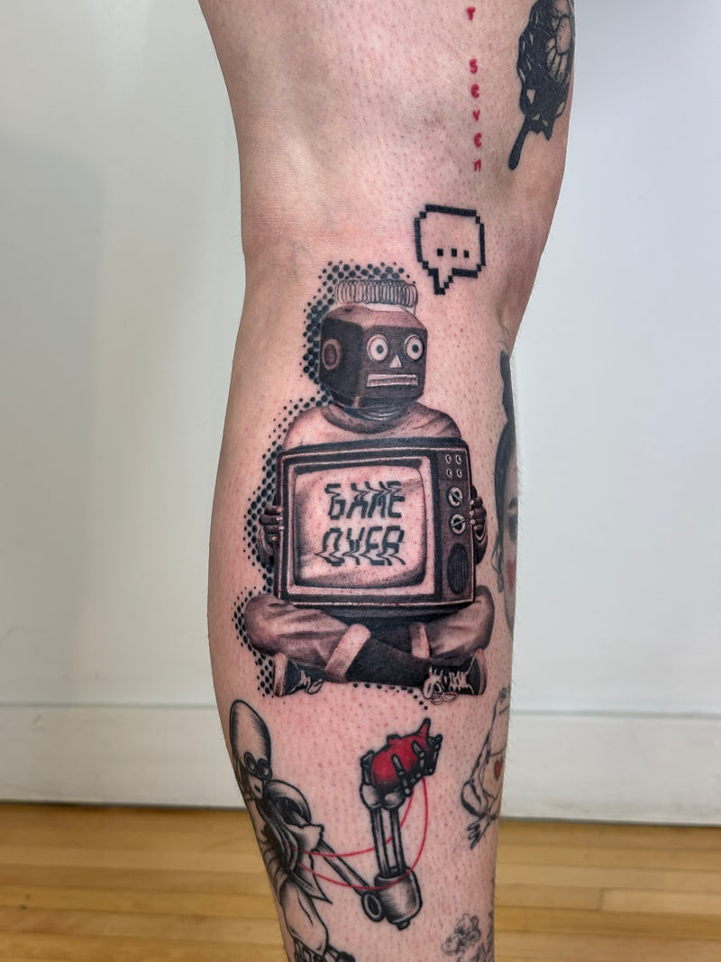 A close-up of a person's leg featuring a detailed black and gray tattoo of a retro robot holding a TV that says 'GAME OVER', with other tattoo designs visible around it.