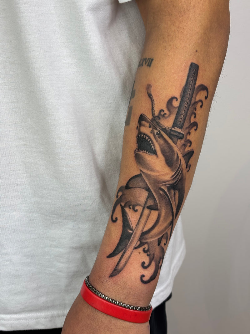 A close-up of a person's forearm featuring a detailed black and gray tattoo of a shark biting into a sword, with tentacle-like swirls. The person is wearing a white t-shirt and a red bracelet.