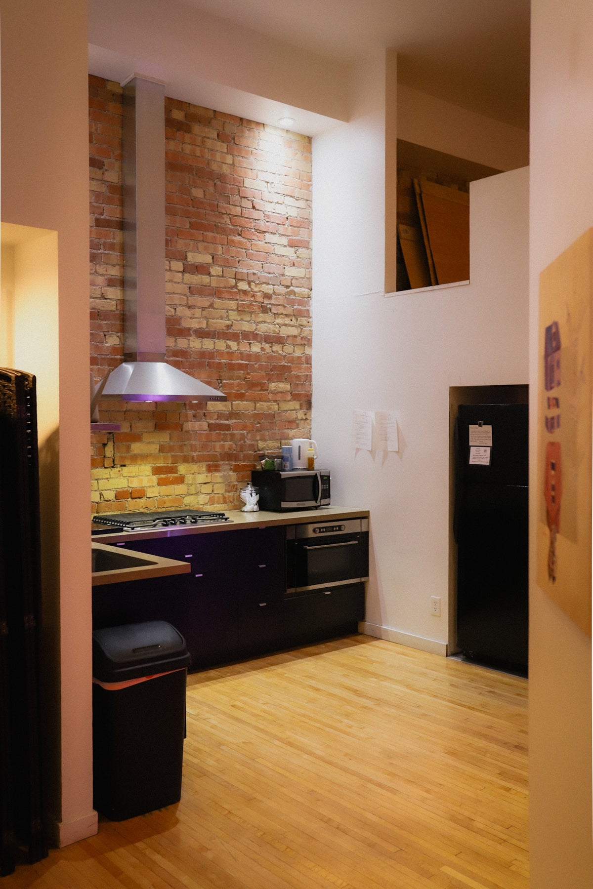 A modern kitchen interior with a dark purple countertop and cabinetry, a brick backsplash, a stainless steel range hood, light wood flooring, and a black refrigerator in the background.