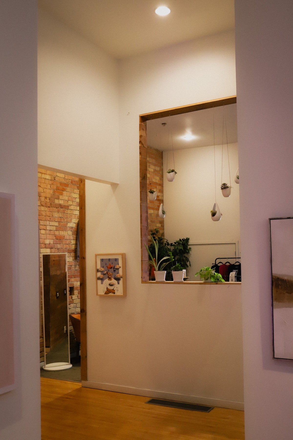 An interior hallway with warm white walls, wood flooring, a brick accent wall, a mirror, and small potted plants on a ledge.