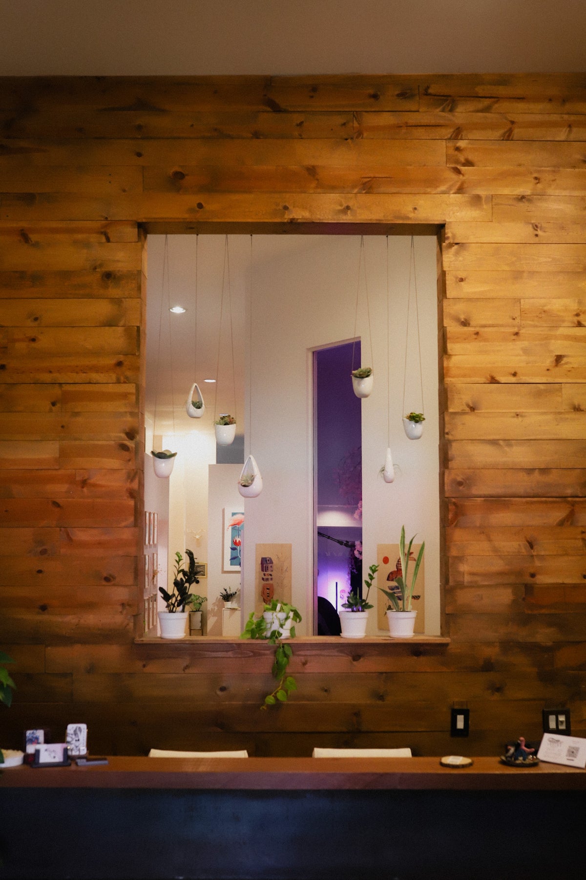 A rustic wooden reception desk set against a warm wood-paneled wall, featuring a large central window looking into the collective tattoo space with hanging plants and warm lighting.