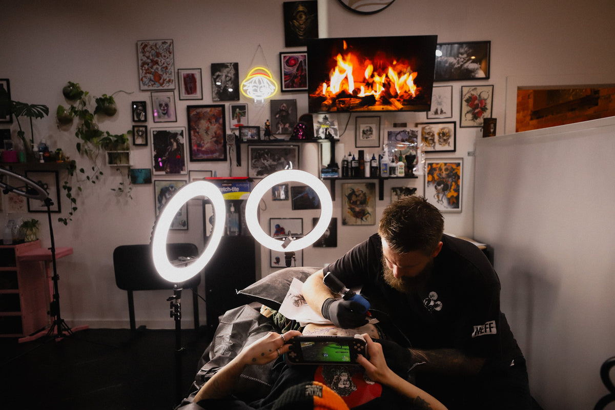 A tattoo artist wearing a black t-shirt with a white graphic on the chest is tattooing a client in a dimly lit room. The scene is illuminated by two bright ring lights and a TV showing a fire, with a wall covered in framed art and posters behind them.