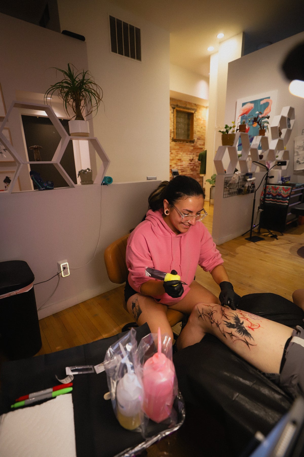 A tattoo artist in a pink hoodie and black gloves applies a tattoo to a person's leg inside a tattoo studio. The artist holds a tattoo needle and a small yellow-and-black tool, with tattoo supplies and the client's tattooed leg visible in the foreground and room background.