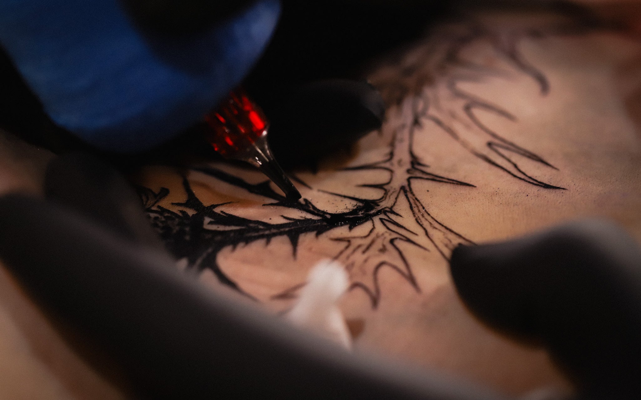A close-up action shot of a tattoo artist's hand holding a tattoo pen, applying black ink to a person's skin in a detailed, spiky tribal pattern.
