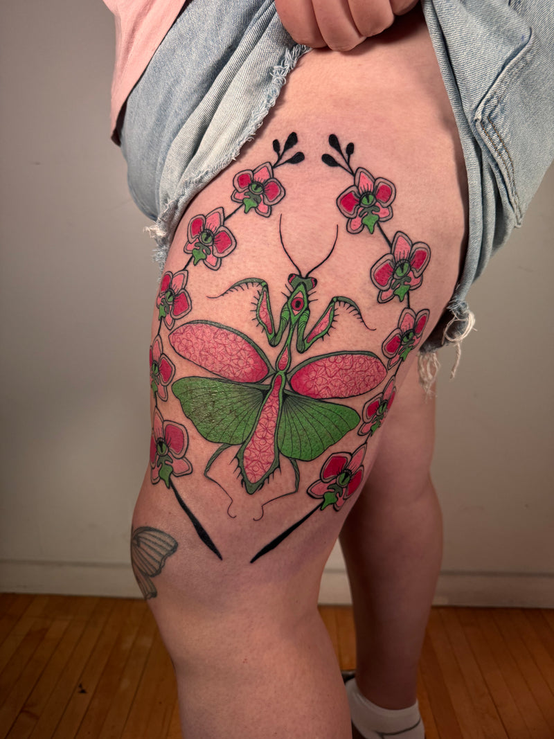 A close-up of a person's upper thigh showing a colorful tattoo of a praying mantis surrounded by pink flowers, with a light denim shirt lifted to reveal the skin and tattoo.