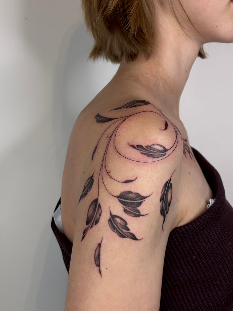 A close-up of a person's shoulder and upper arm featuring a black floral vine and leaf tattoo design, with the person wearing a dark ribbed top against a plain light wall.