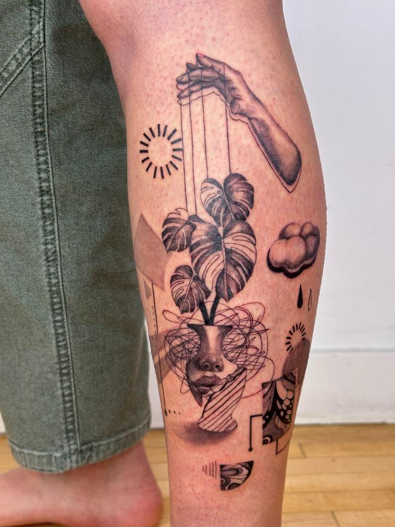 A close-up of a person's calf featuring a detailed black and gray tattoo of a hand puppet with a monstera plant, clouds, and abstract shapes, next to olive green denim jeans.