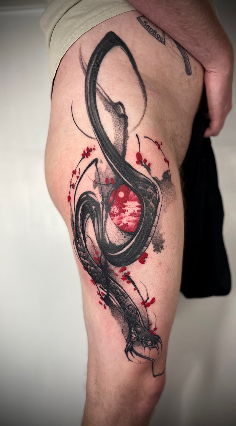 A close-up of a person's upper thigh showing a detailed black and red tattoo design of a stylized snake wrapping around a red sphere, with small red flower accents, against a plain light background.
