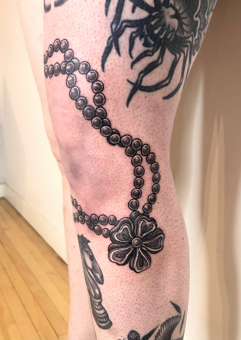 A close-up of a person's leg featuring a detailed black and gray tattoo of a beaded necklace with a flower pendant, alongside other tattoo designs.