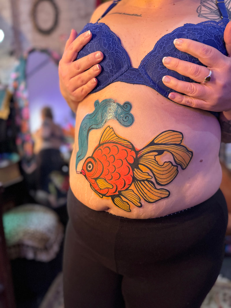 A close-up of a person's midsection showing a colorful temporary tattoo of a koi fish on their stomach, styled with a blue lace bra top and black pants.
