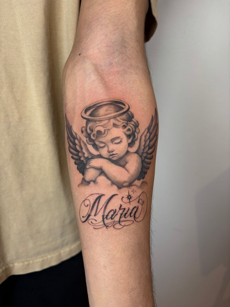 A close-up of a person's forearm featuring a black and gray tattoo of a sleeping cherub with wings, a halo, and the name 'Maria' in cursive script.
