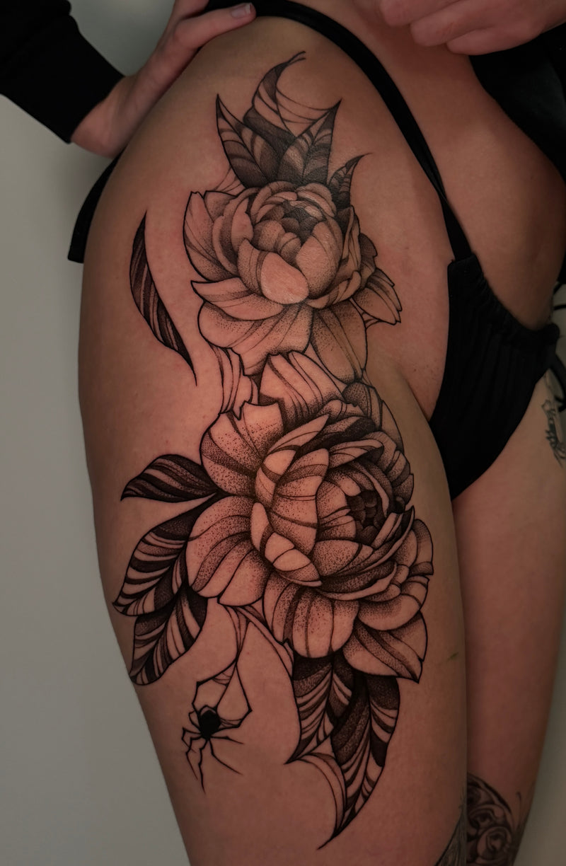 A large black and gray floral tattoo on a person's upper thigh, featuring two layered peony-style flowers with leaves and a small spider detail, shown against skin with black underwear visible.