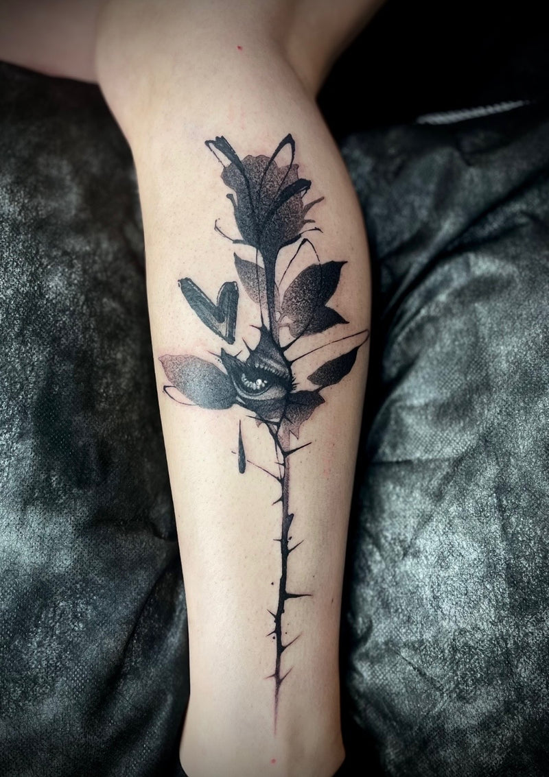 A black and gray watercolor-style tattoo of a rose with thorns on a person's calf, shown against a dark textured fabric background.