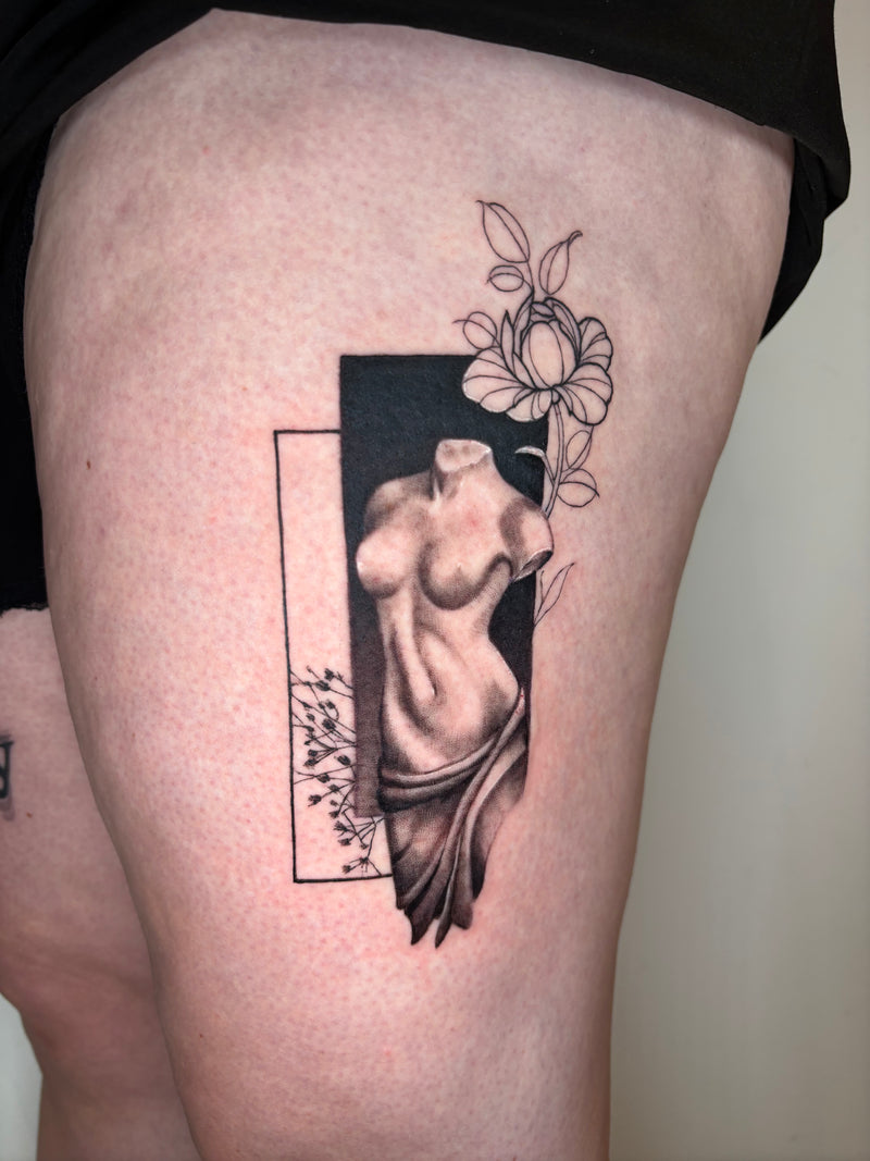 A close-up of a thigh showing a freshly tattooed design. The black and gray tattoo features a classical female nude torso framed by a black rectangle, with a black-and-white flower above and delicate floral details on the left side.