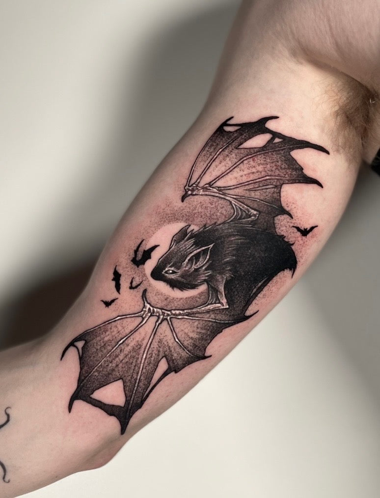 A detailed black and gray tattoo of a bat with spread wings on a person's bicep