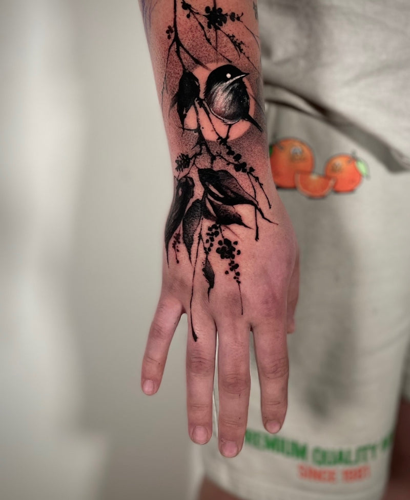 A close-up of a hand and forearm featuring a realistic black ink tattoo of two birds perched on branches with leaves and berries, shown against a light gray background.