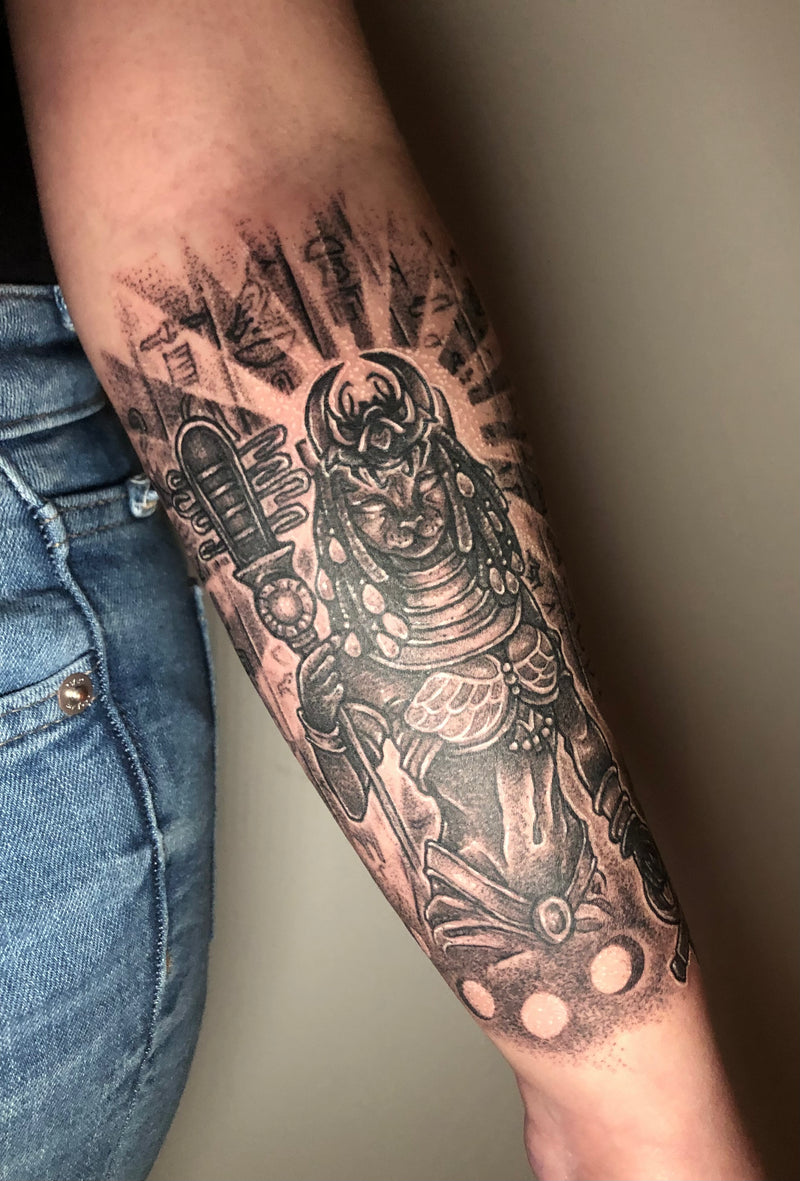 A close-up of a person's forearm featuring a detailed black and gray tattoo of an Egyptian deity holding a flintlock pistol, styled with military insignia and rays behind the figure.
