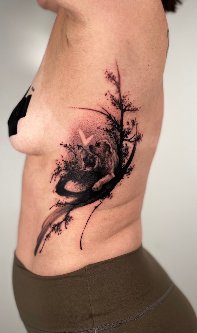 A close-up of a person's side torso showing a detailed black and gray tattoo of a sleeping cat nestled against a dog among tree branches and leaves on the upper hip.