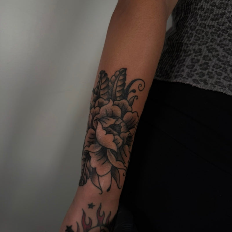 A close-up of a person's forearm featuring a detailed black and gray traditional-style floral tattoo with a large flower and leaves, worn with a gray patterned top and black pants.