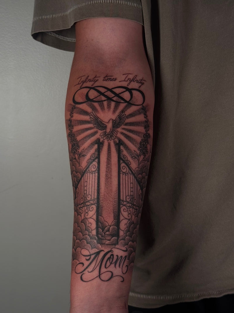 A close-up of a person's forearm featuring a large black and gray tattoo of a gate, a dove, rays, clouds, and the words 'Infinity times Infinity' and 'Moms'.