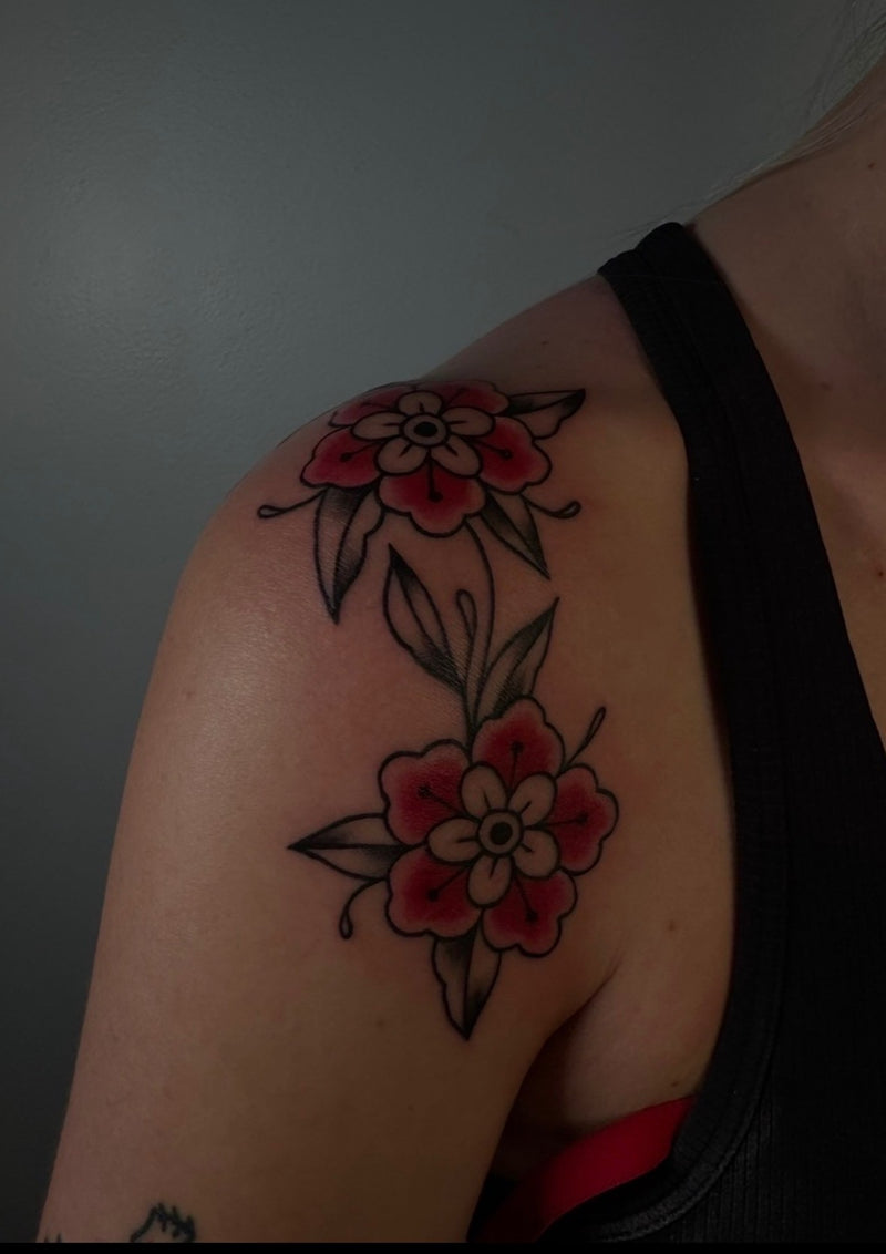 A close-up of a person's upper shoulder and arm featuring a traditional-style floral tattoo with two red and pink flowers, black outlines, and green leaves on the skin.