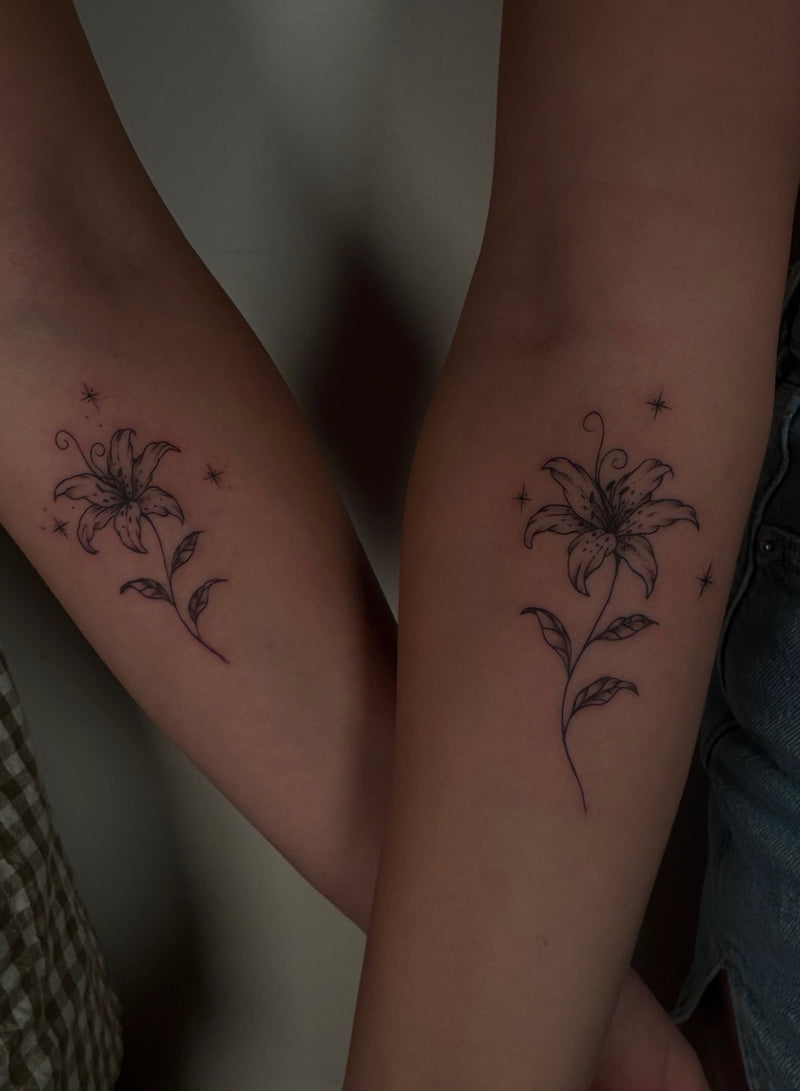 Two forearm tattoos featuring minimalist black lily flowers with stems and small star accents, shown on a person's arms against a dim indoor background.