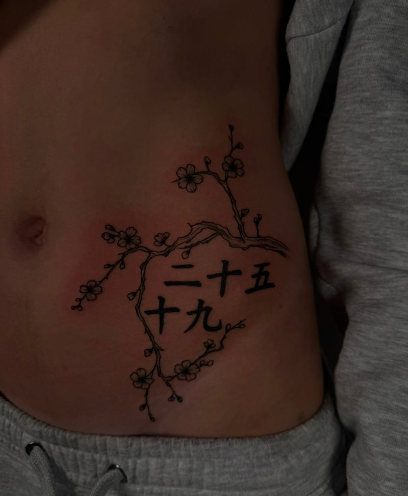 A close-up of a person's side torso showing a black floral branch tattoo with Chinese characters on the abdominal wall, wearing a gray sweatshirt.