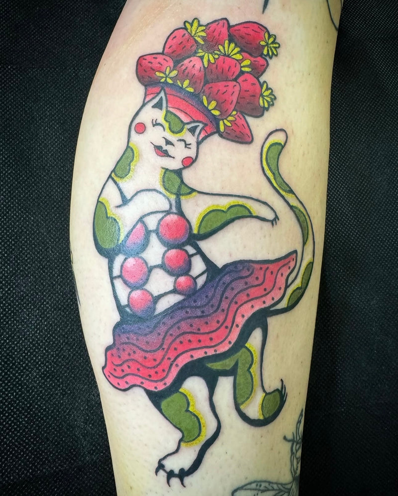 A close-up of a tattoo on human skin depicting a stylized white cat wearing a red strawberry hat and a pink-red skirt, with green and black outlines, set against a dark fabric background.